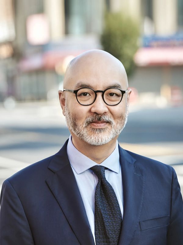 A smiling bald man with a beard wearing glasses and a navy suit outdoors.