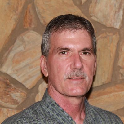 Middle-aged man with a mustache in a checkered shirt against a stone wall.