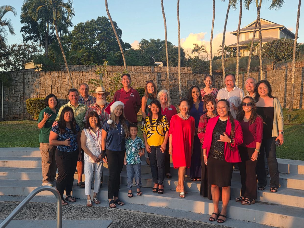 Group of people posing outdoors with palm trees and stone wall.
