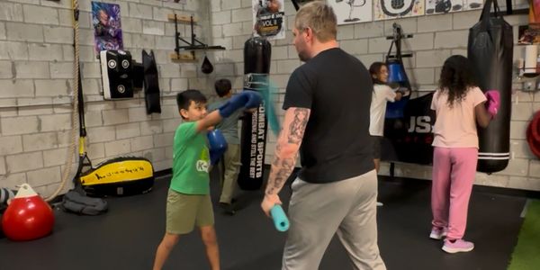 A boxing coach trains children in a gym with punching bags and gloves.