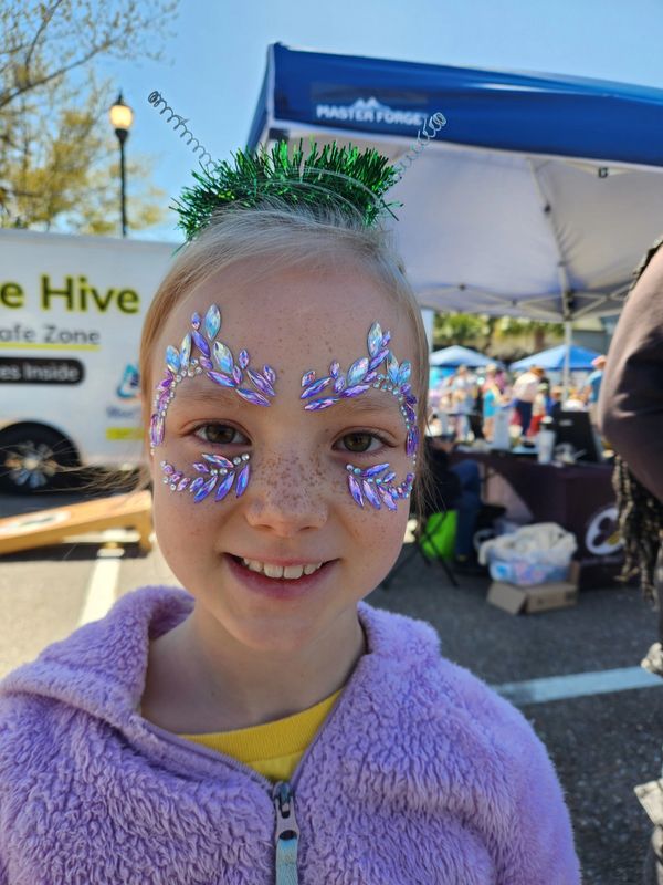 Smiling child with face gems and festive antennae headband at an outdoor event.