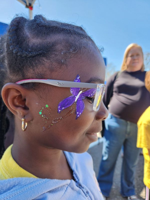 A young girl with glasses and a purple butterfly face paint smiles outdoors.
