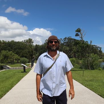 Man wearing sunglasses and a brown hat walking on a sunny park path.
