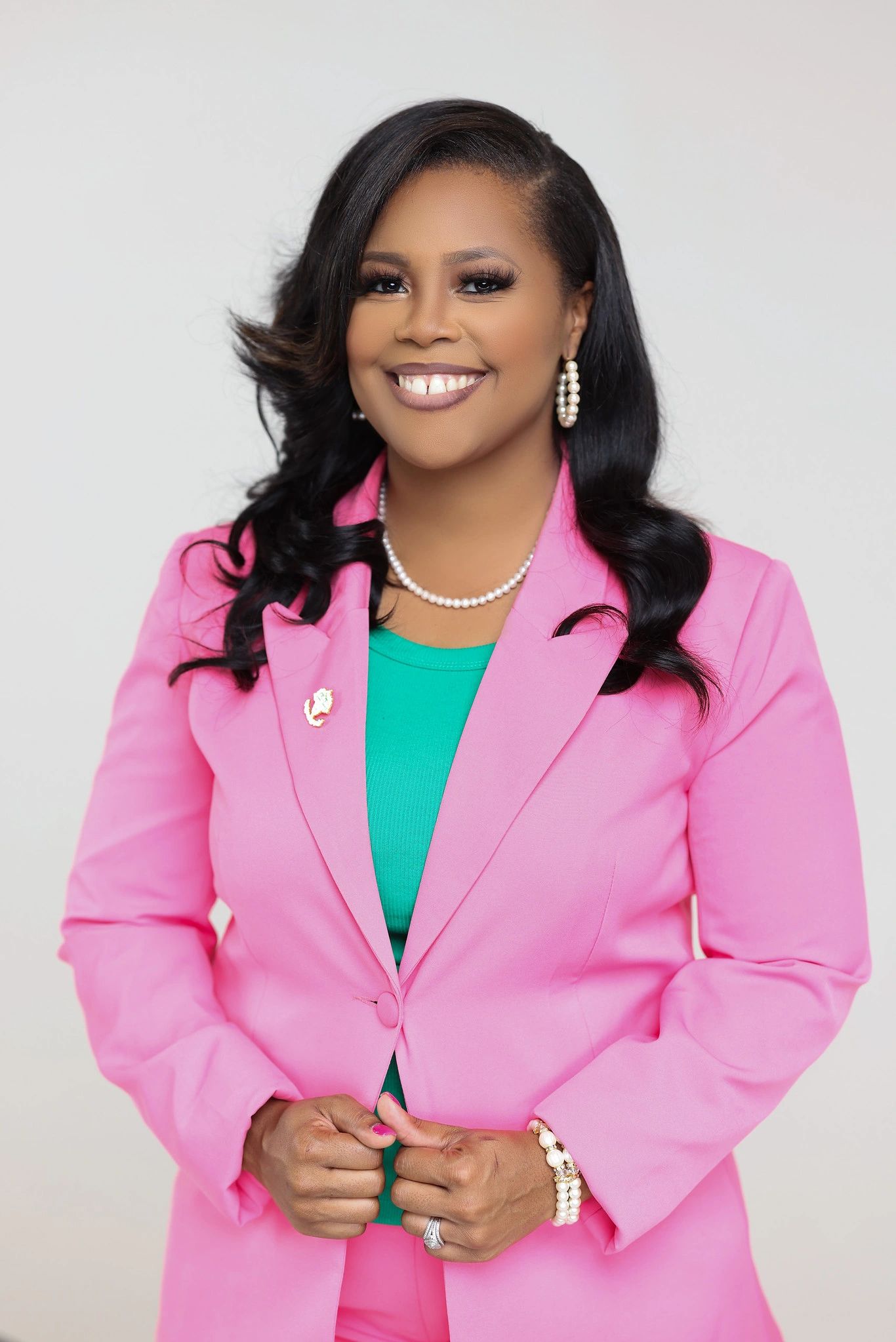Confident woman smiling, dressed in a pink blazer and green top, accessorized with pearl jewelry.
