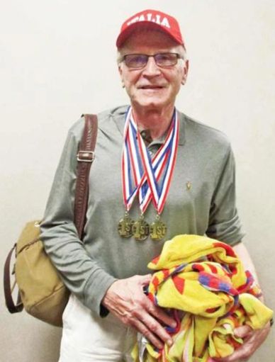 Elderly man wearing medals and holding colorful towels with a bag over his shoulder.