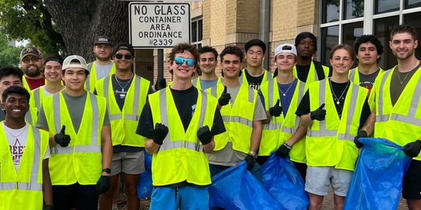 Aggie Business Kings smiling by an Adopt A Street Sign with yellow vests and trash bags.