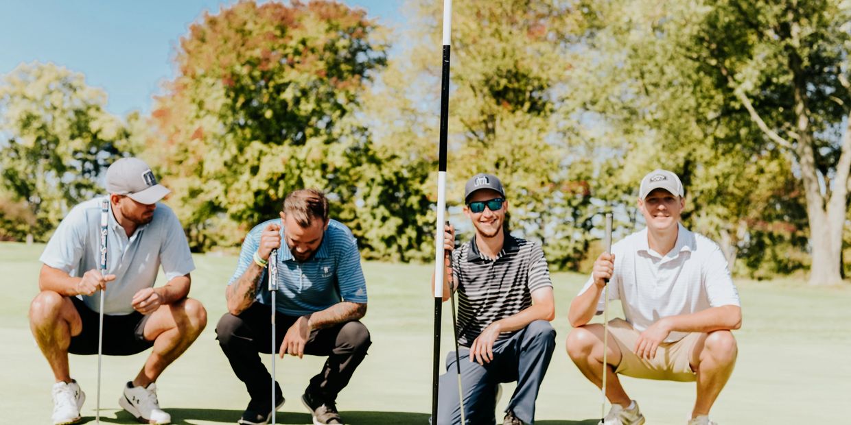 Four men squatting on a golf course holding putters next to a flag.
