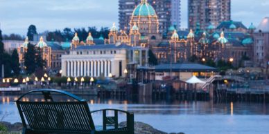 Empty bench overlooking a lit-up cityscape by the water at dusk.