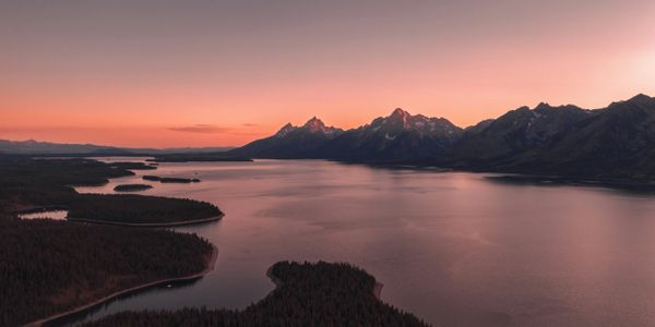 Peaceful lake at sunset with mountain silhouettes and orange sky.