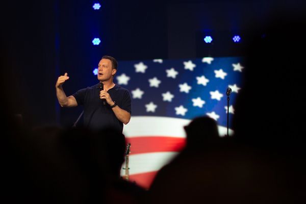 Man speaking on stage with American flag background.