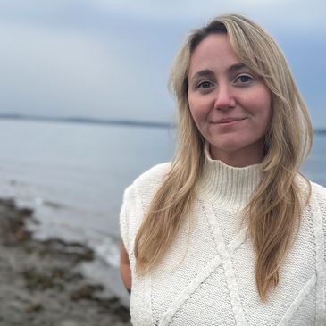 Woman in a white sweater standing by a calm lakeside on a cloudy day.