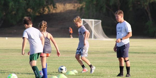 Kids practicing soccer drills on a grassy field with cones and a goal in the background.