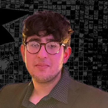A young man with glasses and curly hair wearing a dark blazer.