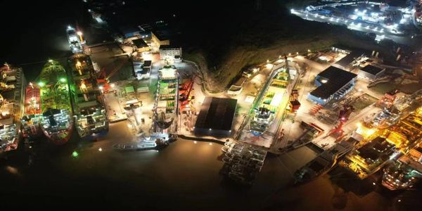 Aerial nighttime view of a brightly lit industrial port with docked ships and infrastructure.