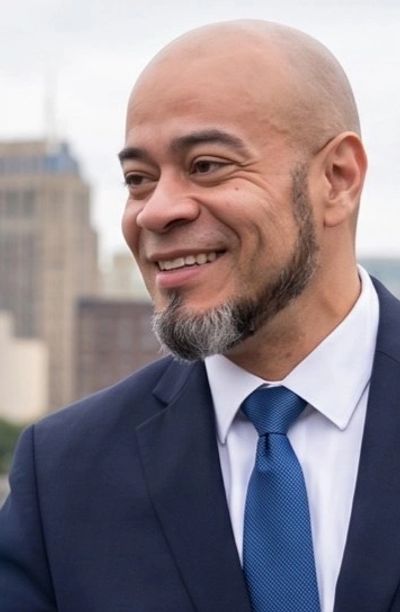 Smiling bald man with a trimmed beard in a blue suit and tie outdoors.