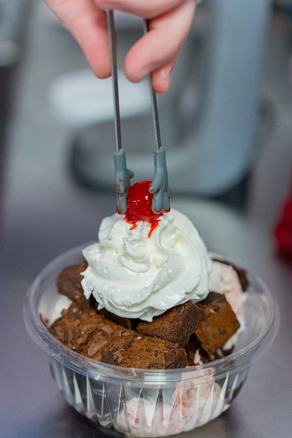 A hand places a cherry on whipped cream topping brownies in a clear cup.