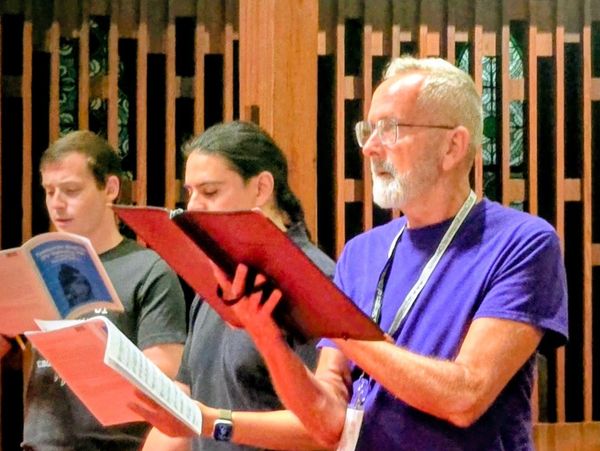 Three men singing from sheet music in a wooden-paneled room.