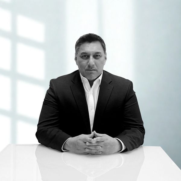 Serious man in a black suit sitting at a white table with hands clasped.