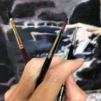 Hand holding three paintbrushes against a blurred painting background.