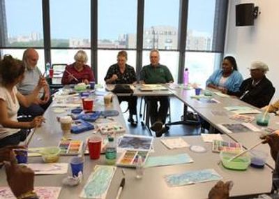 A diverse group of adults participates in a painting class, surrounded by art supplies.