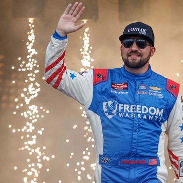 Race car driver in patriotic suit waves with fireworks behind.
