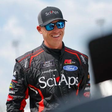 Race car driver in SciAps racing suit and cap smiling outdoors.