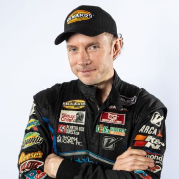 Race car driver in black Menards racing suit and cap.