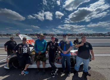 Group of six people standing by a race car on a sunny day at a racetrack.