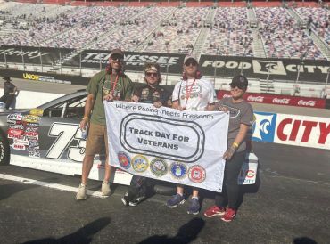 Three people holding a Track Day for Veterans banner at a race track beside a car.