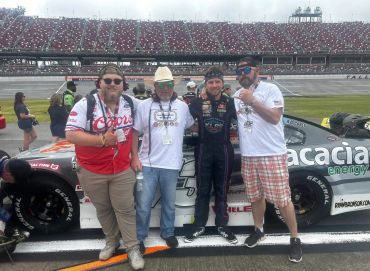 Four men posing in front of a race car at a crowded racetrack.