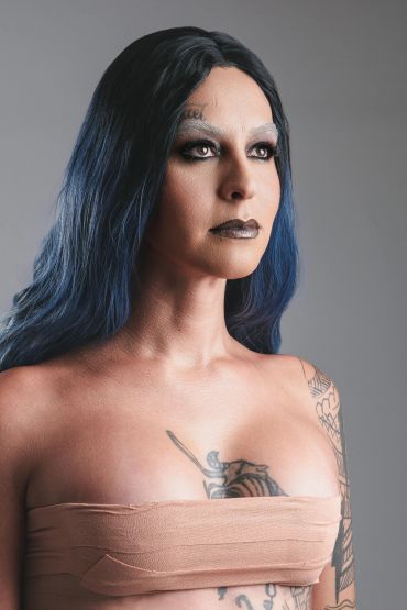 Woman with blue-black hair and tattoos, wearing neutral makeup and a bandage top.