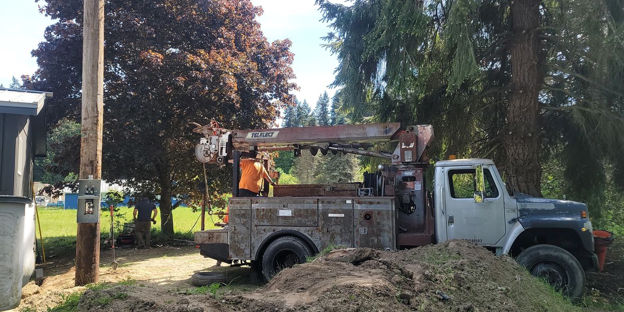 Utility truck with workers near trees and dirt piles on a sunny day.