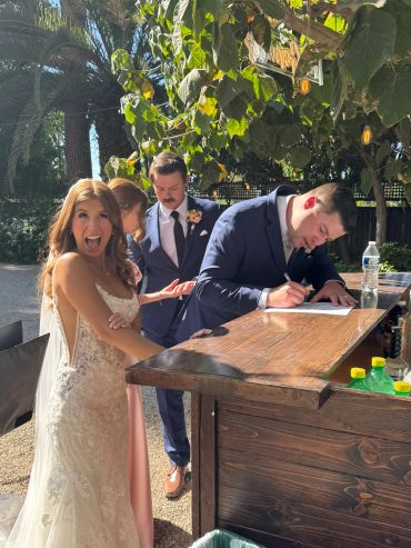 Bride excitedly pointing while groom signs a document outdoors.