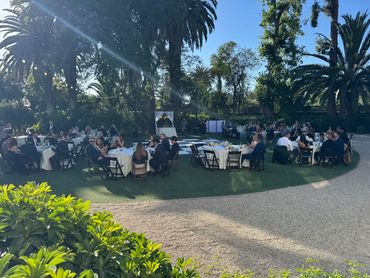 Outdoor formal gathering with round tables and people.