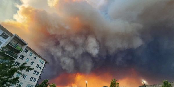 Massive smoke clouds billowing from a wildfire near residential buildings at sunset.