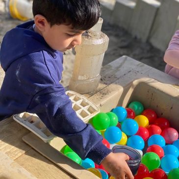 A child playing with colorful plastic balls and a cup in a sandbox.