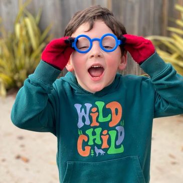 Child in red gloves and blue glasses plays outdoors with a joyful expression.