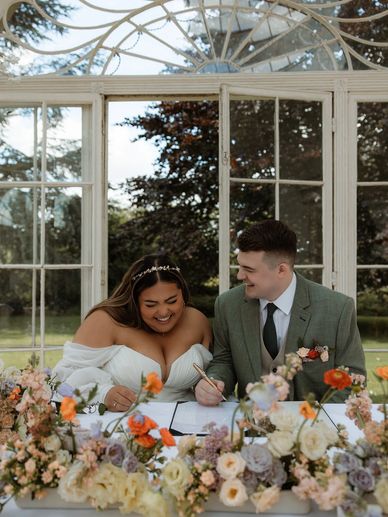 Bride and groom happily signing wedding documents at a floral-decorated table.