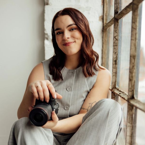 Smiling woman with camera sitting by a rustic window.