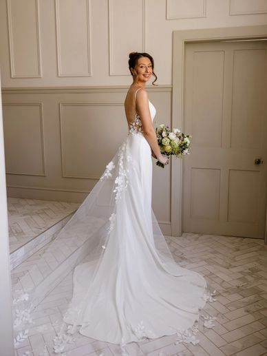 Bride in elegant white wedding dress with floral veil holding bouquet indoors.