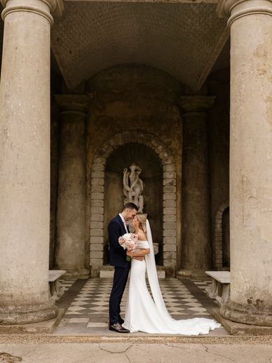 Bride and groom embrace in front of classical stone pillars and statue.