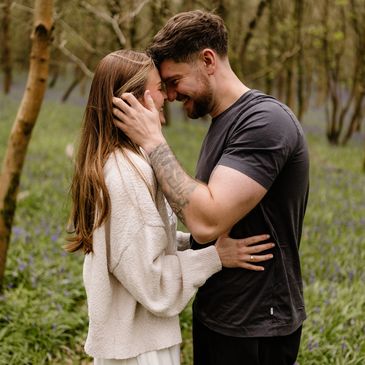 A couple shares an intimate moment in a forest, smiling and touching foreheads.