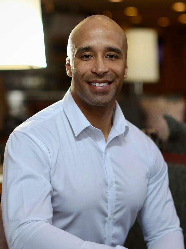 A man seated and smiling. Light brown skin. Bald head. White dress shirt with top button undone. 
