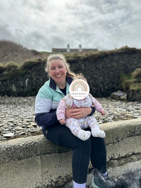 Woman sitting on a stone wall holding a baby, outdoors near a rocky area.