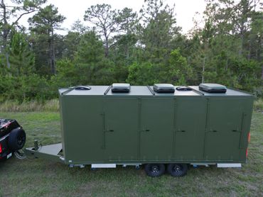 Military green trailer with ventilation units, set in a forest clearing.