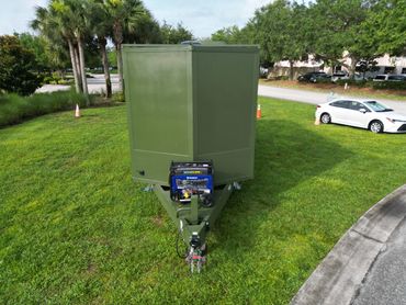 Green trailer with a portable generator parked on grass near a road and palm trees.