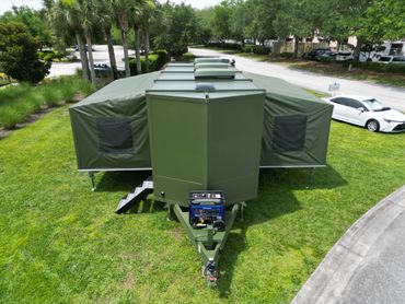 Military-style green tent trailer set up on grassy area near palm trees and parked cars.