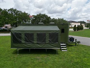 Olive green military trailer with an extended canopy and steps on a grassy field.