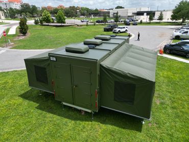 Military-style green mobile unit with extended sides on grass near a parking area.