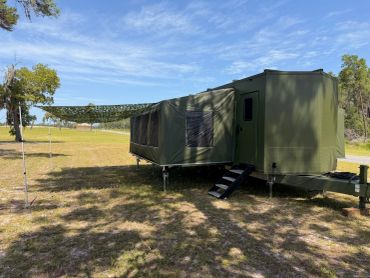 Military-style green trailer with camouflage shade on a grassy field.
